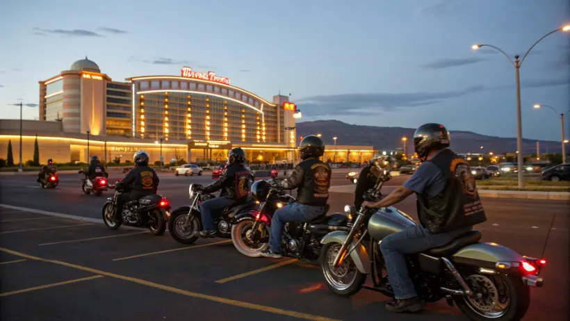 A group of motorcyclists in leather vests and helmets parking their bikes in front of a large illuminated casino resort entrance at dusk