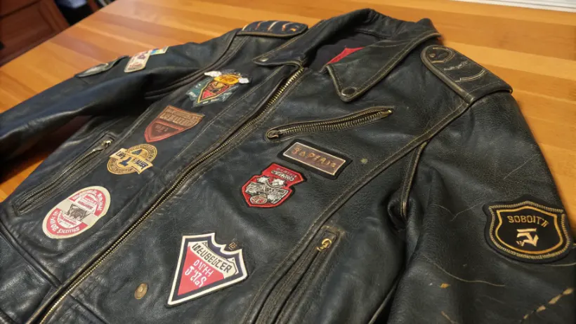 Close-up of a worn black leather biker jacket covered in embroidered club patches and road pins, laid flat on a wooden surface